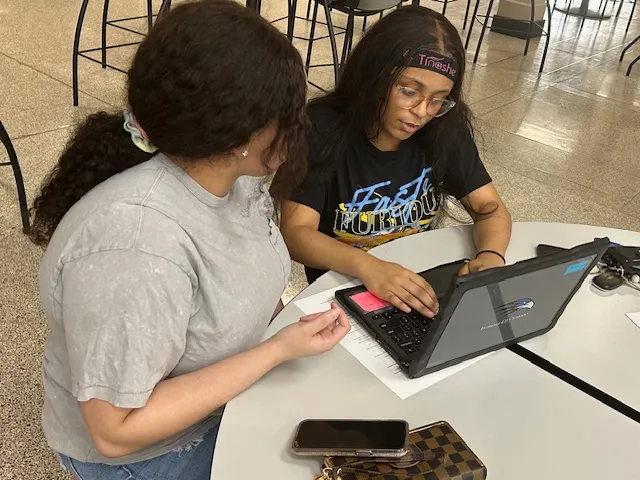 Two people sit at a table. The person on the right is using a laptop